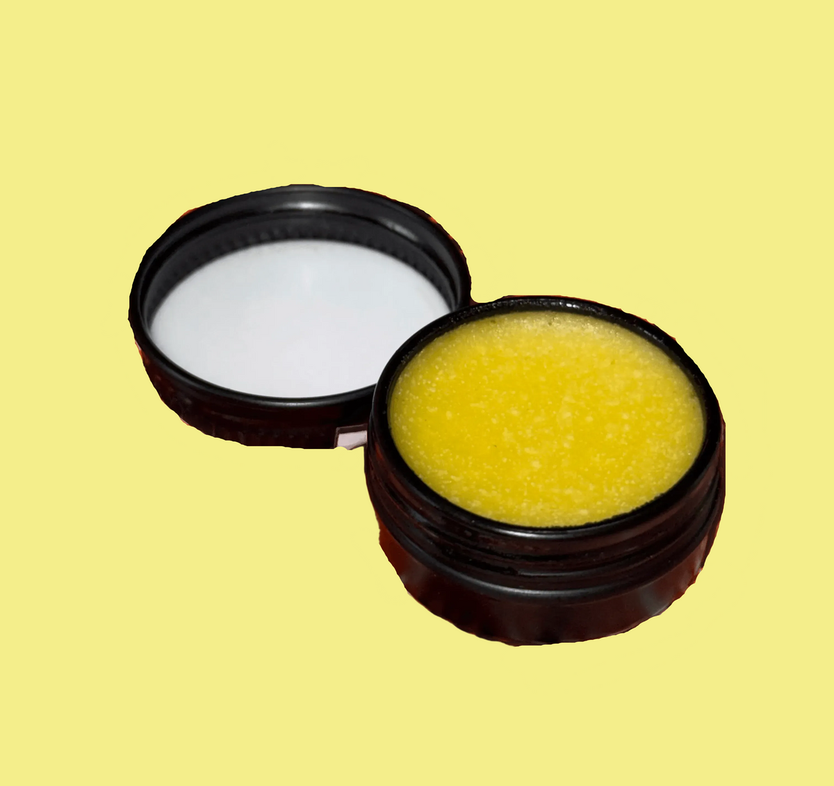 A small round black container shown open, with the lid placed beside it. The container holds a bright yellow balm or cream, photographed on a plain yellow background.