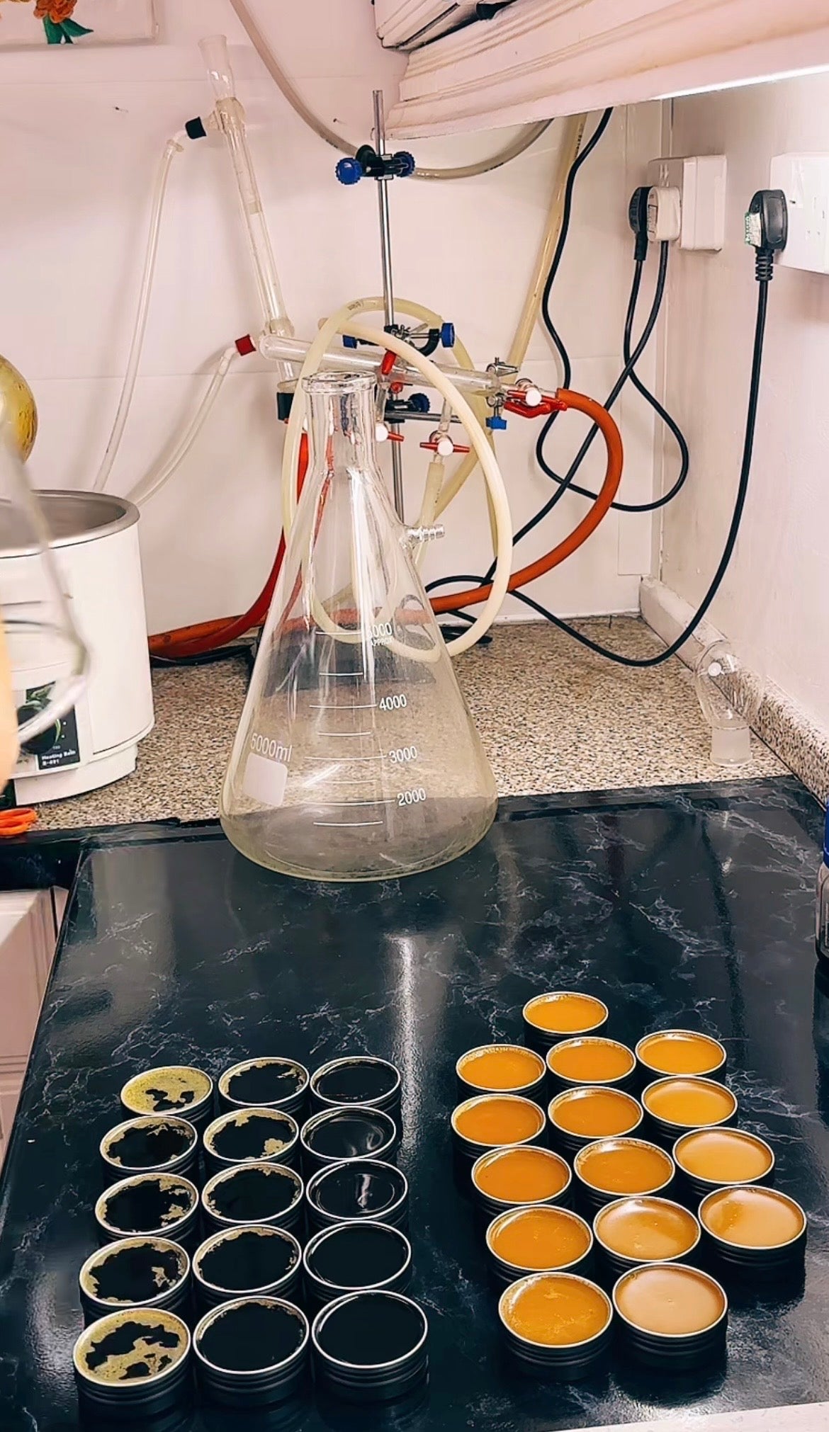 A clear glass conical flask sits on a dark countertop in a kitchen or workshop setting, with several small round metal tins containing dark and golden-yellow substances arranged nearby. White walls, electrical outlets, and tubing are visible in the background.