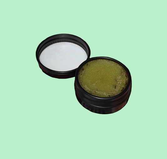 An open round black metal jar showing a textured yellow-green balm or ointment, with its black lid placed beside it on a plain light green background.