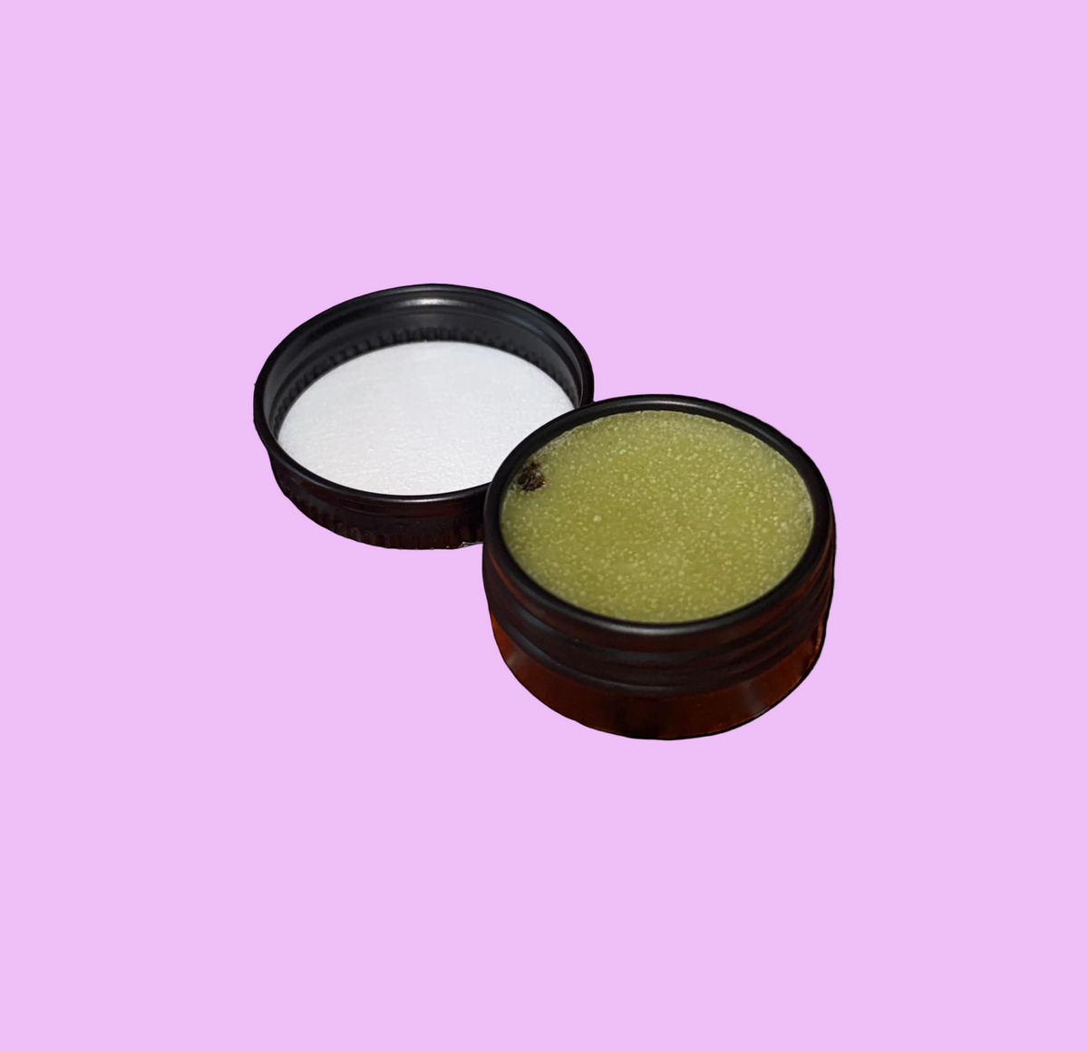 An open round black metal jar showing a textured green balm or ointment, with its black lid placed beside it on a plain light purple background.