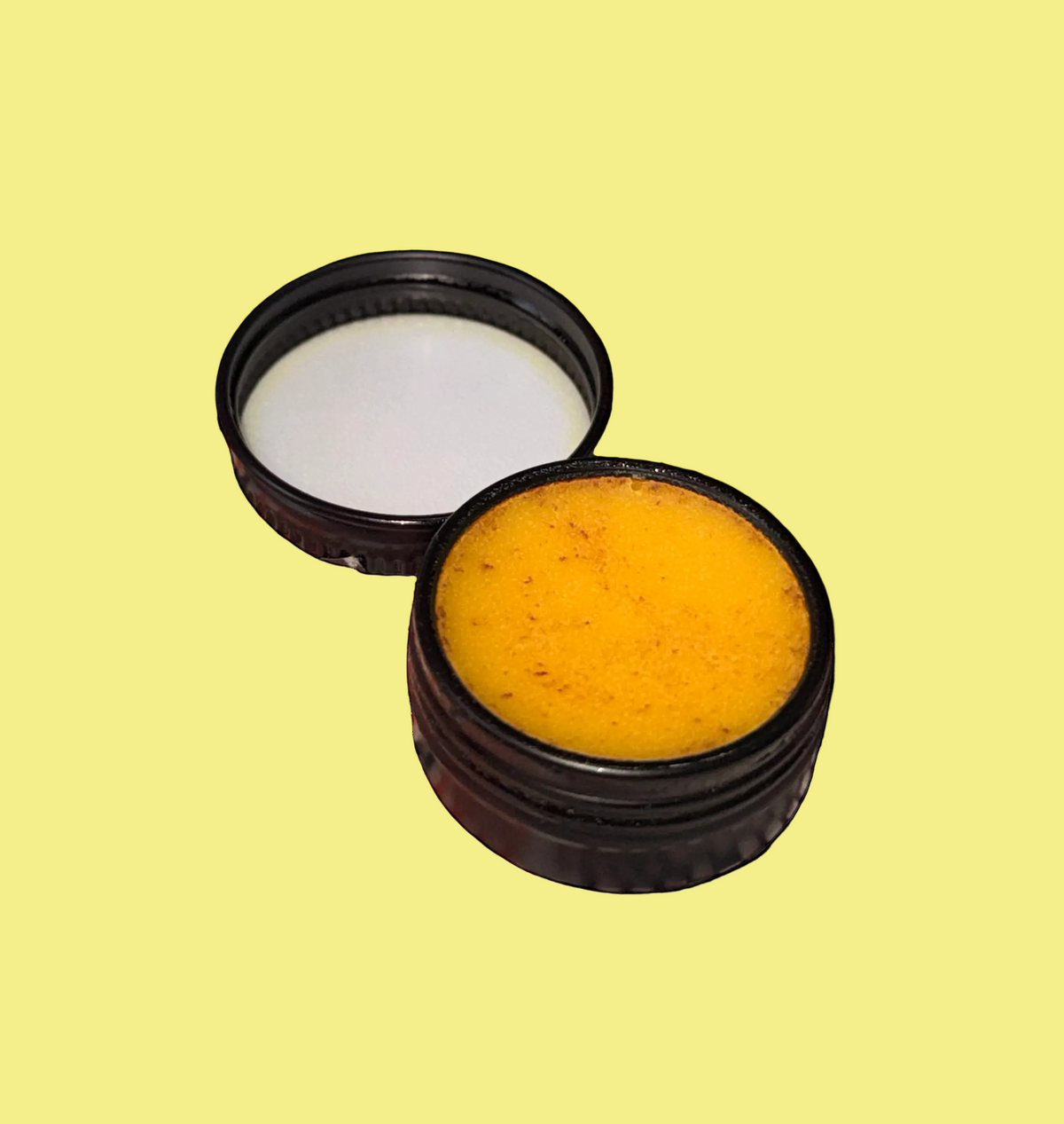 An open round black metal jar showing a textured orange with small red flecks balm or ointment, with its black lid placed beside it on a plain light yellow-orange background.