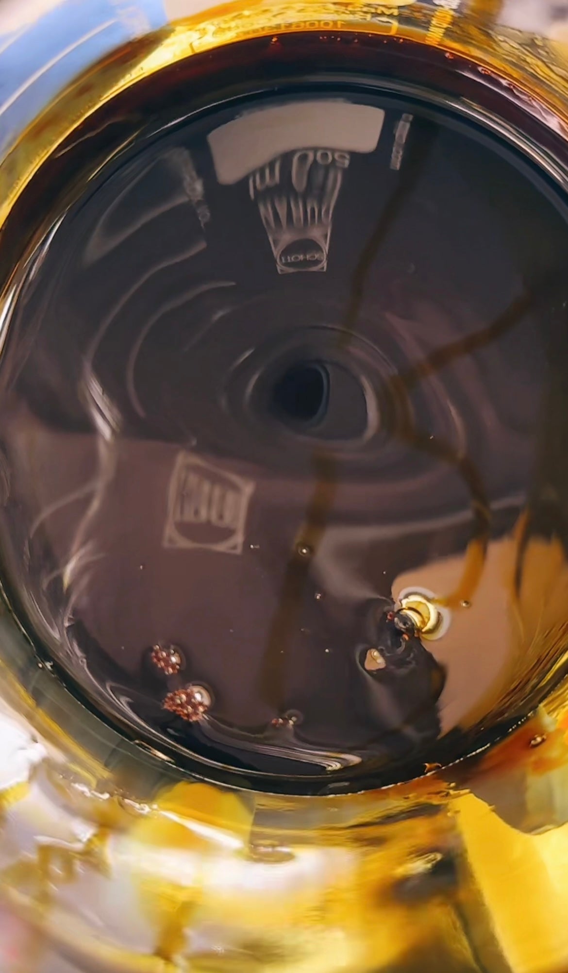 Close-up of a clear glass bottle or container filled with dark amber ginger extract, showing the base and reflective surface with small bubbles and a faint printed label near the top.