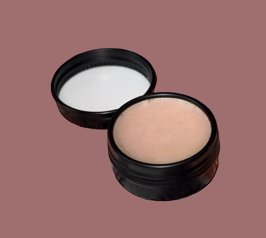 An open round black container showing a pale pink cream or balm, with the matching black lid placed behind it on a plain salmon background.
