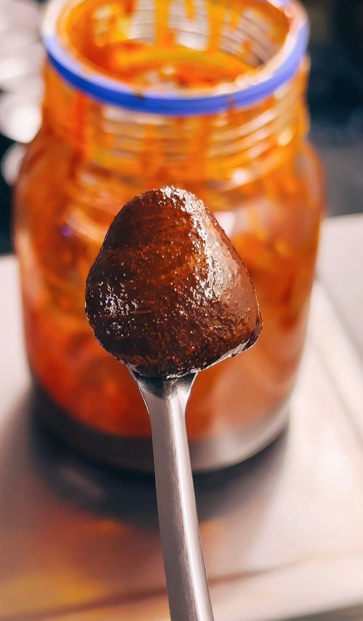 A close-up shot of a glossy, dark reddish-brown tumeric and black pepper extract held on a metal spoon in front of an open glass jar with orange-red contents.